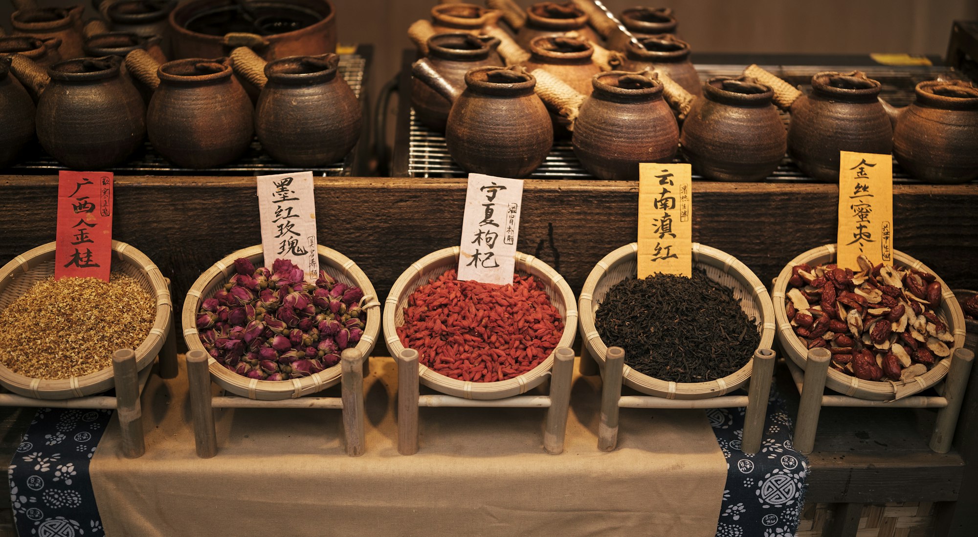 Woven bamboo baskets of dried osmanthus, rosebuds, goji berries, tea and red dates with handwritten Chinese labels, in front of clay herb pots