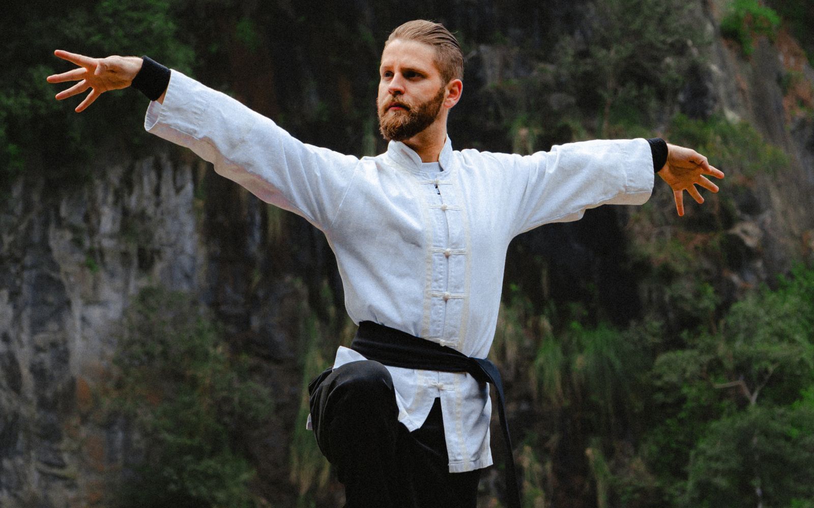 A man in a white kung fu jacket practising tai chi in front of a forested cliff